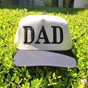 May include: White baseball cap with a black brim and the word "DAD" in black letters on the front.