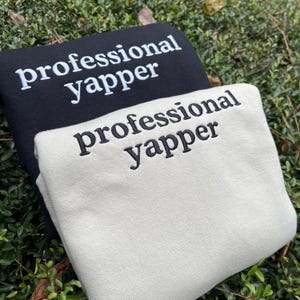 May include: Two sweatshirts with the text "professional yapper" embroidered on them. The top sweatshirt is black with white text, and the bottom sweatshirt is white with black text.