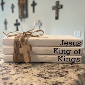 May include: A stack of three white wooden blocks with the words "Jesus King of Kings" written in black. The blocks are tied together with twine and a small wooden cross is attached to the twine.