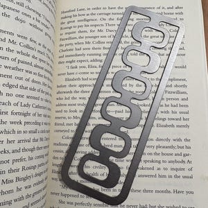 May include: A silver metal bookmark with a unique, abstract design featuring a series of interconnected, rounded shapes. The bookmark is placed on an open book with text visible.