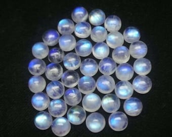 Puede incluir: Colección de cabujones redondos de piedra lunar pulida. Cada piedra muestra una base blanca lechosa con adularescencia azul, creando un brillo suave y etéreo. Las piedras están dispuestas juntas sobre un fondo oscuro.