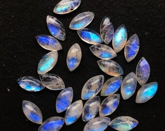 Puede incluir: Una colección de piedras preciosas de piedra lunar de forma ovalada con un brillo nacarado. Las piedras muestran una gama de colores, incluyendo blanco, azul y toques de iridiscencia. Las piedras preciosas están dispersas sobre una superficie oscura.