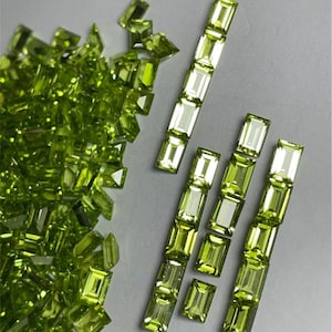 May include: A collection of faceted peridot gemstones in various shapes. Some are scattered, while others are arranged in vertical rows. The gems have a vibrant, light green color and a rectangular or emerald cut.