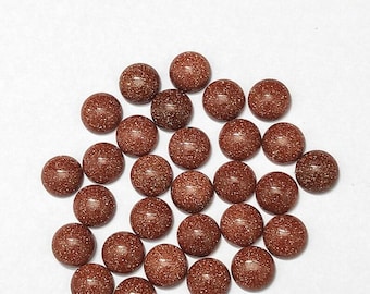 May include: A collection of round, polished, brown gemstones with a speckled, glittery appearance. The gemstones are scattered on a white surface. Each stone is approximately 0.5 inches in diameter.