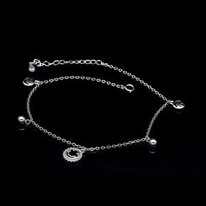 Puede incluir: Una delicada tobillera plateada con una serie de pequeños colgantes. La tobillera presenta un colgante de carita sonriente rodeado de pequeñas piedras transparentes, junto con varias pequeñas cuentas plateadas y colgantes de carita sonriente. La tobillera se muestra sobre un fondo negro.