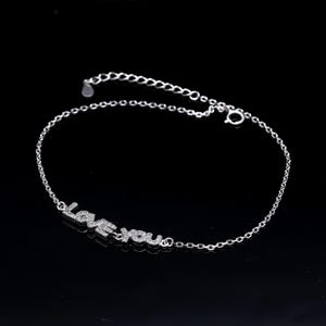Puede incluir: Una pulsera plateada con las palabras "LOVE YOU" escritas con piedras preciosas claras y brillantes. La pulsera tiene una cadena delicada y un cierre seguro. La joya se muestra sobre un fondo negro.