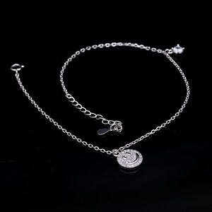 Puede incluir: Un collar de plata con un colgante de carita sonriente. El colgante redondo está incrustado con pequeñas piedras transparentes. La cadena del collar es delicada y ajustable. Una pequeña gema transparente está al final de la cadena.