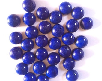 May include: A collection of polished, deep blue lapis lazuli cabochons. The round gemstones are scattered across a white surface, each displaying a smooth, reflective surface. The stones vary slightly in size and have a subtle, natural marbling.