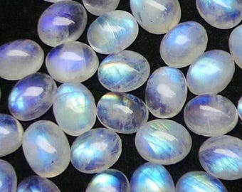May include: A collection of oval-shaped moonstone cabochons. The stones display a milky white base color with iridescent blue and rainbow flashes. The gemstones are arranged closely together against a dark background, showcasing their natural beauty and potential for jewelry making.