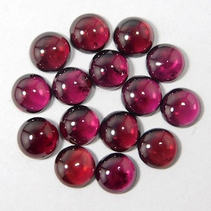 Pode incluir: Uma coleção de quatorze pedras preciosas redondas, lapidadas em cabochão. As gemas são de uma cor vermelho-púrpura profunda e rica, com um acabamento polido e brilhante. As pedras estão dispostas em um padrão circular sobre um fundo branco.