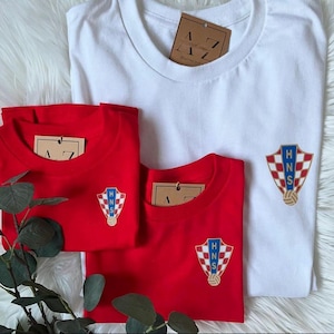 May include: Three t-shirts, one white and two red, with a Croatian national team crest embroidered on the front. The crest features a checkered shield with the letters "HNS" in gold.