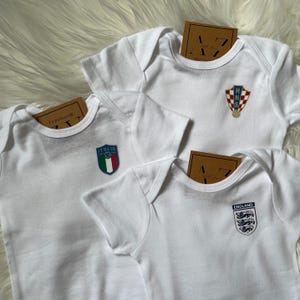 May include: Three white baby onesies with different country flags printed on them. The flags are for Italy, Croatia, and England.