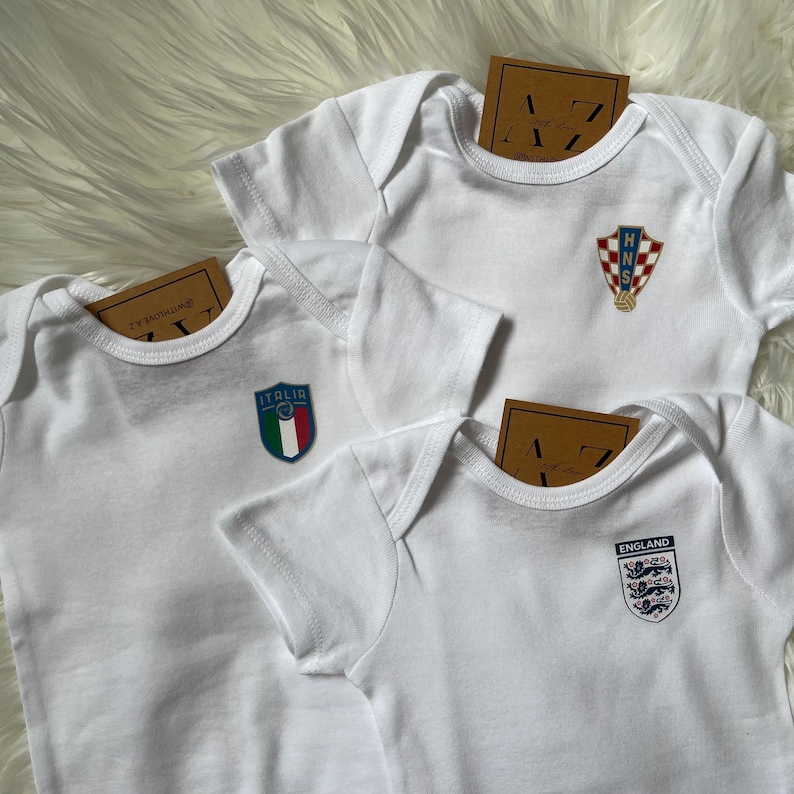 May include: Three white baby onesies with different country flags printed on them. The flags are for Italy, Croatia, and England.