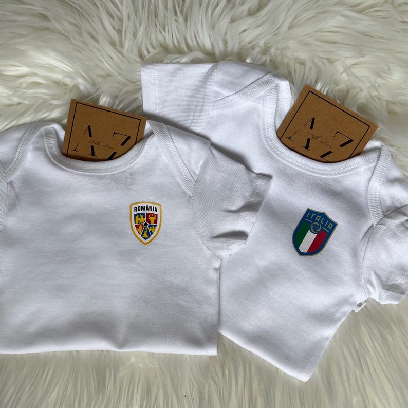 May include: Two white baby onesies with embroidered patches. One onesie has a patch with the words "Romania" and a coat of arms. The other onesie has a patch with the words "Italia" and the Italian flag.
