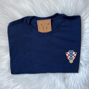 May include: A navy blue t-shirt with a gold and red Croatian national team crest on the left chest. The crest features the letters "HNS" and a soccer ball.