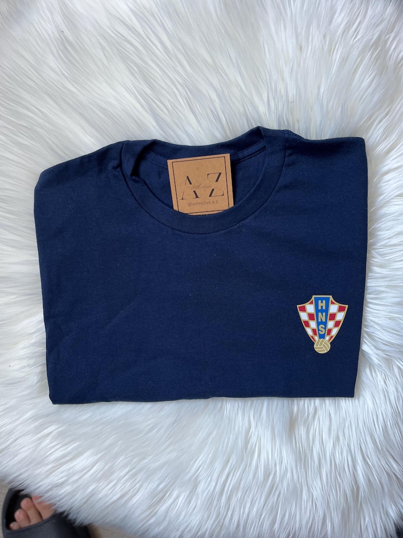 May include: A navy blue t-shirt with a gold and red Croatian national team crest on the left chest. The crest features the letters "HNS" and a soccer ball.