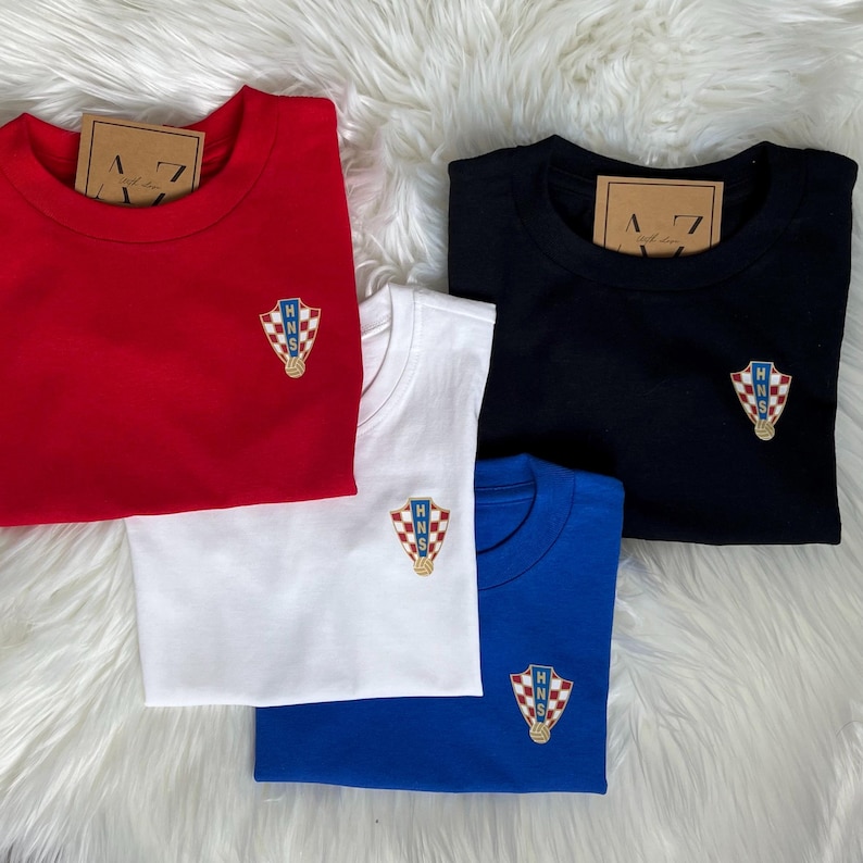 May include: Four t-shirts in red, white, black, and blue with a Croatian national team logo embroidered on each. The logo features a red and white checkered shield with the letters "HNS" in blue.