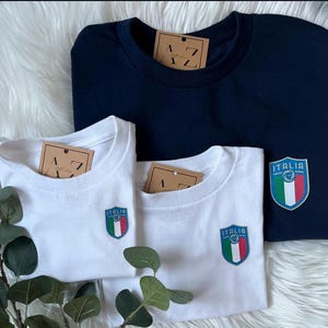 May include: Two white t-shirts with a blue and green Italian flag patch and the word "ITALIA" on the front. A navy blue t-shirt is stacked on top of the white t-shirts.