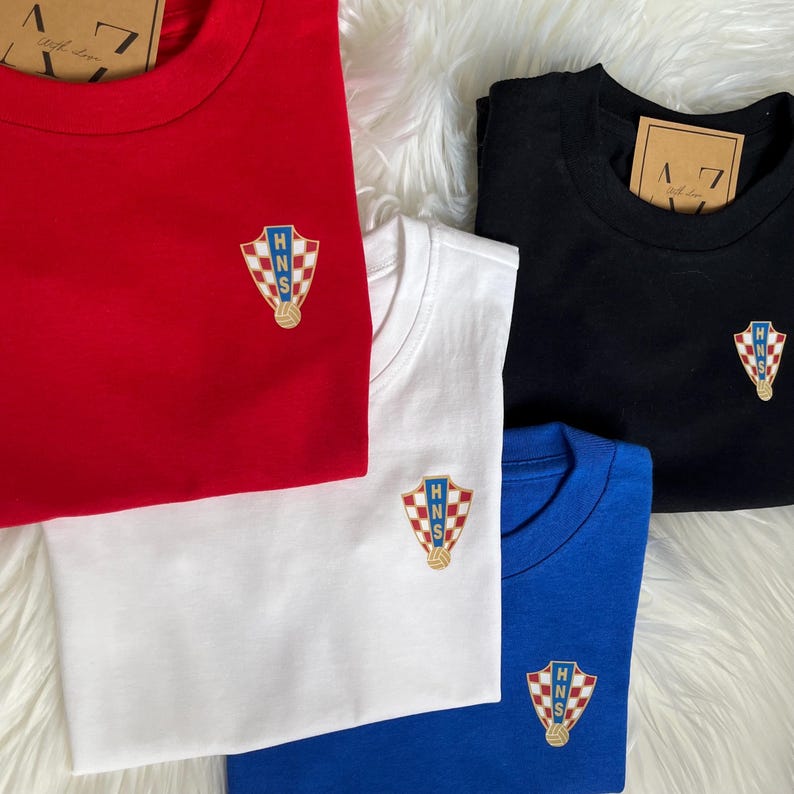 May include: Four t-shirts in red, white, black, and blue with a Croatian national team logo embroidered on the front. The logo features a checkered shield with the letters "HNS" inside.