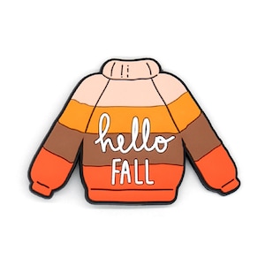 K&ouml;nnte beinhalten: Eine pulloverf&ouml;rmige Anstecknadel mit horizontalen Streifen in den Farben Hellrosa, Gelb, Braun und Orange. Der Text "hello FALL" ist in wei&szlig;er Schreib- und Blockschrift in der Mitte des kleidungsst&uuml;ckf&ouml;rmigen Accessoires zu sehen.