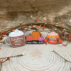 K&ouml;nnte beinhalten: Drei Emaille-Anstecknadeln mit Herbstmotiven. Eine ist eine braune Tasse mit wei&szlig;em Schaum und dem Text Pumpkin Spice and everything nice. Eine weitere zeigt drei K&uuml;rbisse mit dem Text happy FALL y'all. Die dritte ist ein Pullover mit dem Text hello FALL.