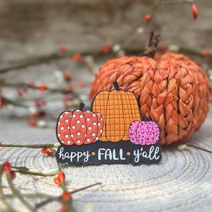 K&ouml;nnte beinhalten: Ein dekoratives flaches Ornament mit drei K&uuml;rbissen in Orange, Rot mit wei&szlig;en Punkten und Pink mit Leopardenmuster, platziert auf einer schwarzen Basis mit dem wei&szlig;en Schriftzug "happy FALL y'all" in Schreib- und Blockschrift.