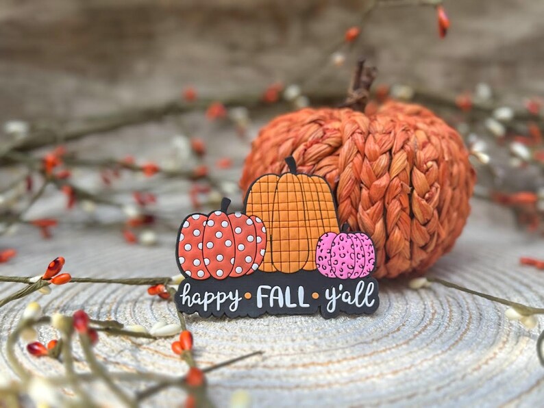 K&ouml;nnte beinhalten: Ein dekoratives flaches Ornament mit drei K&uuml;rbissen in Orange, Rot mit wei&szlig;en Punkten und Pink mit Leopardenmuster, platziert auf einer schwarzen Basis mit dem wei&szlig;en Schriftzug "happy FALL y'all" in Schreib- und Blockschrift.