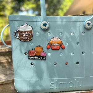 K&ouml;nnte beinhalten: Eine hellblaue Tragetasche aus Gummi mit zwei Griffen und schildkr&ouml;tenf&ouml;rmigen Nieten, verziert mit drei dekorativen Anh&auml;ngern: einer Tasse mit der Aufschrift Pumpkin Spice and everything nice, einem Pullover mit hello FALL und K&uuml;rbissen mit happy FALL y'all.
