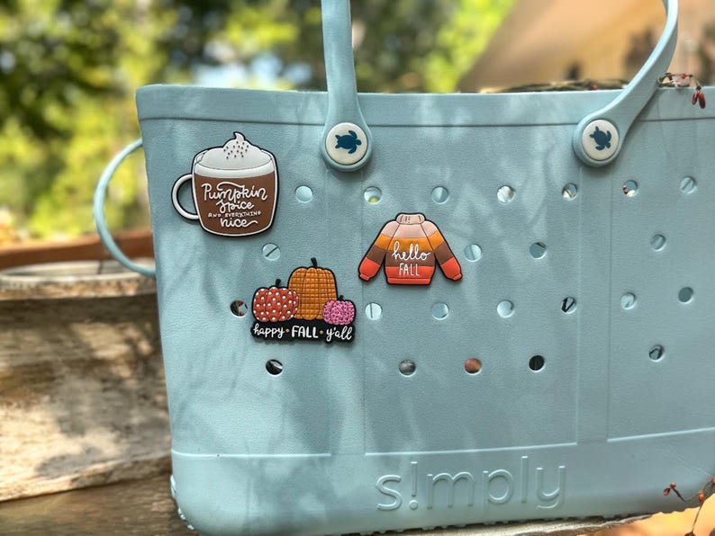 K&ouml;nnte beinhalten: Eine hellblaue Tragetasche aus Gummi mit zwei Griffen und schildkr&ouml;tenf&ouml;rmigen Nieten, verziert mit drei dekorativen Anh&auml;ngern: einer Tasse mit der Aufschrift Pumpkin Spice and everything nice, einem Pullover mit hello FALL und K&uuml;rbissen mit happy FALL y'all.