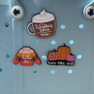 K&ouml;nnte beinhalten: Eine hellblaue Tragetasche aus Gummi mit drei dekorativen Ansteckern. Einer zeigt eine braune Tasse mit wei&szlig;em Schaum und dem Text Pumpkin Spice and everything nice. Ein weiterer ist ein gestreifter Pullover mit dem Text hello FALL. Der dritte zeigt drei K&uuml;rbisse mit dem Text happy FALL y'all.