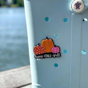 K&ouml;nnte beinhalten: Ein hellblauer Clog-Schuh mit einem dekorativen Anstecker, der drei K&uuml;rbisse in Orange, Rot und Pink mit Punkten und Leopardenmuster zeigt. Der Anstecker enth&auml;lt den wei&szlig;en Schriftzug "happy FALL y'all" auf schwarzem Grund und einen Schildkr&ouml;tenknopf.