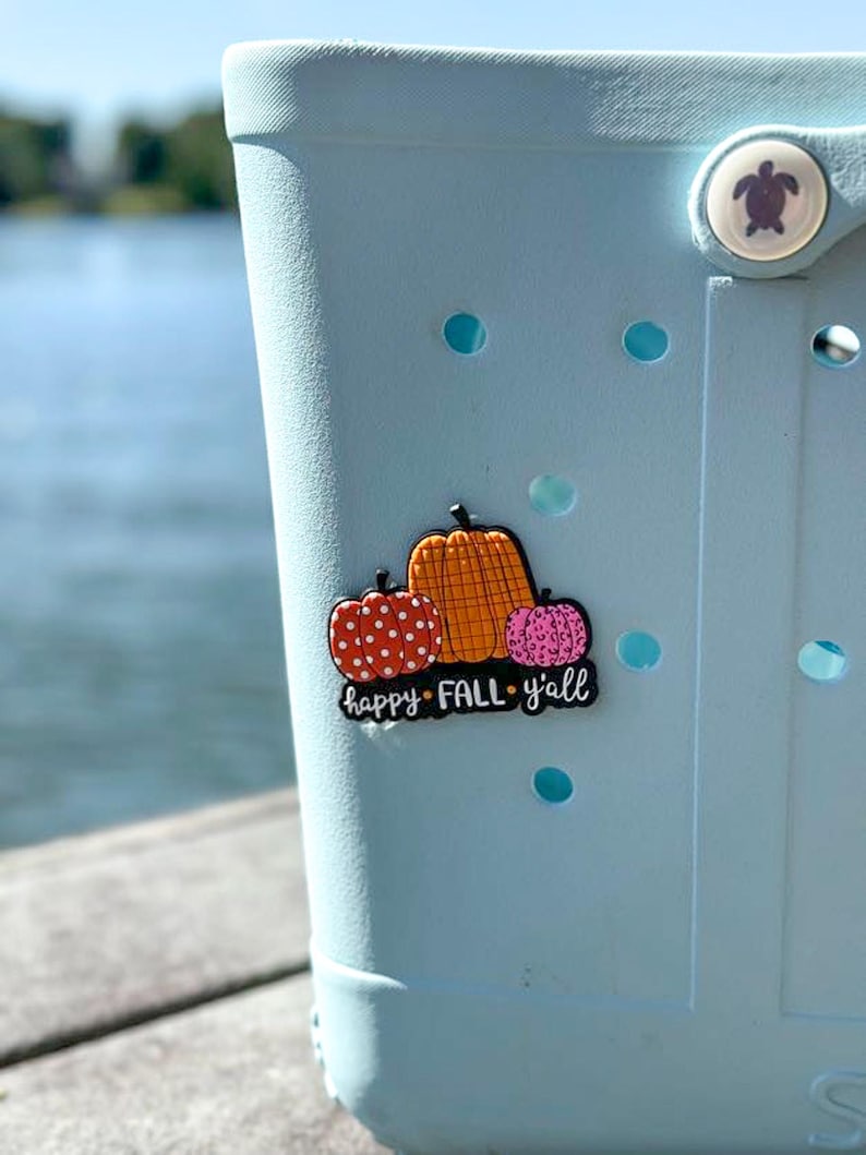 K&ouml;nnte beinhalten: Ein hellblauer Clog-Schuh mit einem dekorativen Anstecker, der drei K&uuml;rbisse in Orange, Rot und Pink mit Punkten und Leopardenmuster zeigt. Der Anstecker enth&auml;lt den wei&szlig;en Schriftzug "happy FALL y'all" auf schwarzem Grund und einen Schildkr&ouml;tenknopf.