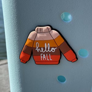 K&ouml;nnte beinhalten: Ein Schuh-Anstecker in Form eines Pullovers mit horizontalen Streifen in Creme, Gelb, Braun und Orange. Der Text hello FALL steht in Wei&szlig; in der Mitte des Pullovers. Der Anstecker hat einen schwarzen Rand und steckt in einem hellblauen Schuh.