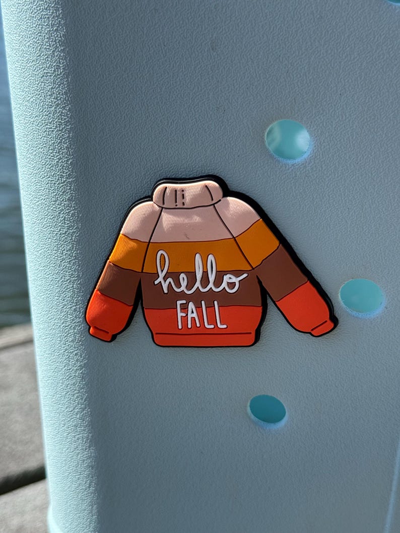 K&ouml;nnte beinhalten: Ein Schuh-Anstecker in Form eines Pullovers mit horizontalen Streifen in Creme, Gelb, Braun und Orange. Der Text hello FALL steht in Wei&szlig; in der Mitte des Pullovers. Der Anstecker hat einen schwarzen Rand und steckt in einem hellblauen Schuh.