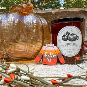 K&ouml;nnte beinhalten: Ein transparenter bernsteinfarbener Glask&uuml;rbis, eine zylindrische Kerze mit schwarzem Deckel und einem Etikett mit der Aufschrift Pumpkin Chai Spice sowie eine pulloverf&ouml;rmige Anstecknadel mit horizontalen Streifen in Rosa, Orange und Braun.
