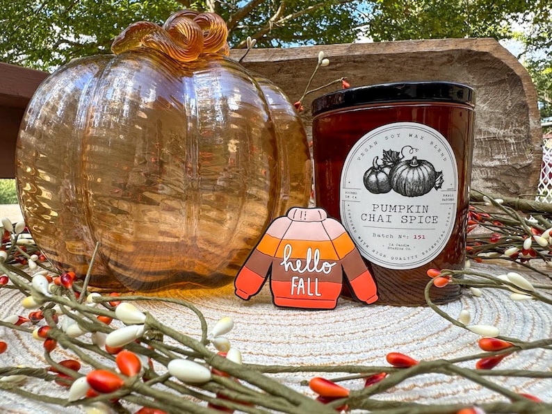 K&ouml;nnte beinhalten: Ein transparenter bernsteinfarbener Glask&uuml;rbis, eine zylindrische Kerze mit schwarzem Deckel und einem Etikett mit der Aufschrift Pumpkin Chai Spice sowie eine pulloverf&ouml;rmige Anstecknadel mit horizontalen Streifen in Rosa, Orange und Braun.