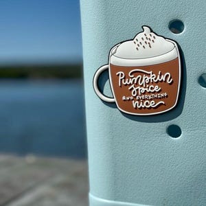 K&ouml;nnte beinhalten: Ein Schuh-Anstecker in Form einer braunen Tasse, gef&uuml;llt mit einem Getr&auml;nk, das mit wei&szlig;er Schlagsahne und braunen Streuseln bedeckt ist. Die Tasse tr&auml;gt den wei&szlig;en Schriftzug Pumpkin Spice AND EVERYTHING nice. Der Anstecker steckt in einem hellblauen Schuh.