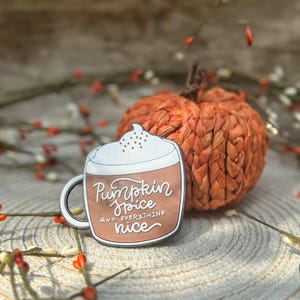 K&ouml;nnte beinhalten: Ein Metall-Emaille-Pin in Form einer Kaffeetasse mit wei&szlig;em Schaum und braunem K&ouml;rper. Die Tasse tr&auml;gt den wei&szlig;en Schriftzug Pumpkin Spice AND EVERYTHING nice. Der Pin liegt neben einer kleinen, strukturierten, orangefarbenen K&uuml;rbisdekoration.