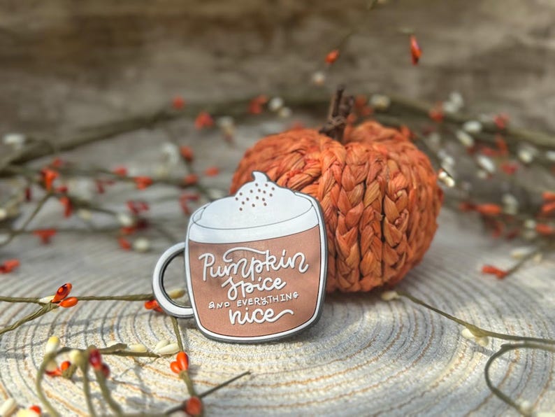 K&ouml;nnte beinhalten: Ein Metall-Emaille-Pin in Form einer Kaffeetasse mit wei&szlig;em Schaum und braunem K&ouml;rper. Die Tasse tr&auml;gt den wei&szlig;en Schriftzug Pumpkin Spice AND EVERYTHING nice. Der Pin liegt neben einer kleinen, strukturierten, orangefarbenen K&uuml;rbisdekoration.