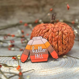 K&ouml;nnte beinhalten: Ein bunter Emaille-Pin in Form eines Pullovers mit horizontalen Streifen in den Farben Rosa, Orange, Braun und Rot. In der Mitte des Pullovers befindet sich der wei&szlig;e Schriftzug "hello FALL" in einer Kombination aus Schreib- und Druckschrift.