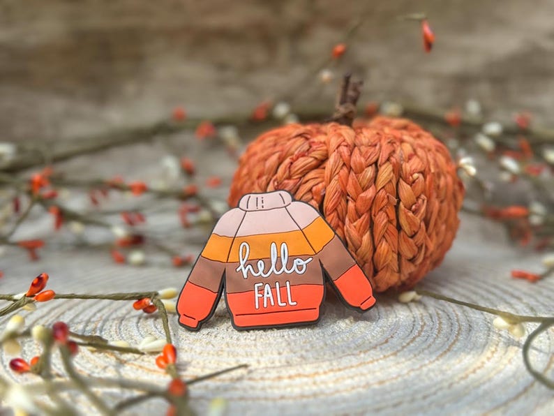K&ouml;nnte beinhalten: Ein bunter Emaille-Pin in Form eines Pullovers mit horizontalen Streifen in den Farben Rosa, Orange, Braun und Rot. In der Mitte des Pullovers befindet sich der wei&szlig;e Schriftzug "hello FALL" in einer Kombination aus Schreib- und Druckschrift.