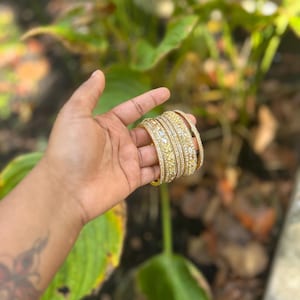 May include: A set of gold bangles with intricate designs and embedded gemstones. The bangles are held in a hand, showcasing their ornate details and the warm, golden hue of the metal. The jewelry is set against a blurred background of green foliage.