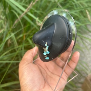 Puede incluir: Un pendiente de color dorado con piedras turquesas colgantes. El pendiente se exhibe sobre un objeto negro y redondeado, sostenido por una mano. El fondo presenta hierba verde.