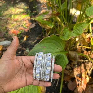 Puede incluir: Un conjunto de pulseras plateadas con piedras cuadradas transparentes y azul claro. Las pulseras se sostienen en una mano, sobre un fondo de follaje verde y hojas marrones otoñales.