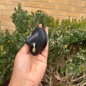 Pode incluir: Um porta-brincos preto em forma de gota com uma corrente dourada. Um brinco pendurado com pedras preciosas azuis, vermelhas e verdes está preso ao suporte. O suporte é segurado em uma mão contra um fundo de folhagem verde e uma parede de tijolos.