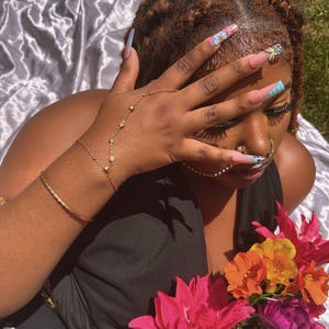 May include: A hand with a gold bracelet, a hand chain, and long, colourful nails. The nails display pink, blue, and floral designs. A bouquet of pink, orange, and purple flowers is in the foreground. The person is wearing a black top.