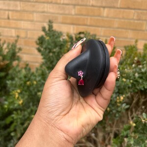 May include: A pair of pink and red gemstone earrings. The earrings are held by a black object. The earrings have a floral design with a teardrop-shaped red gemstone hanging below. The background is a brick wall and green foliage.