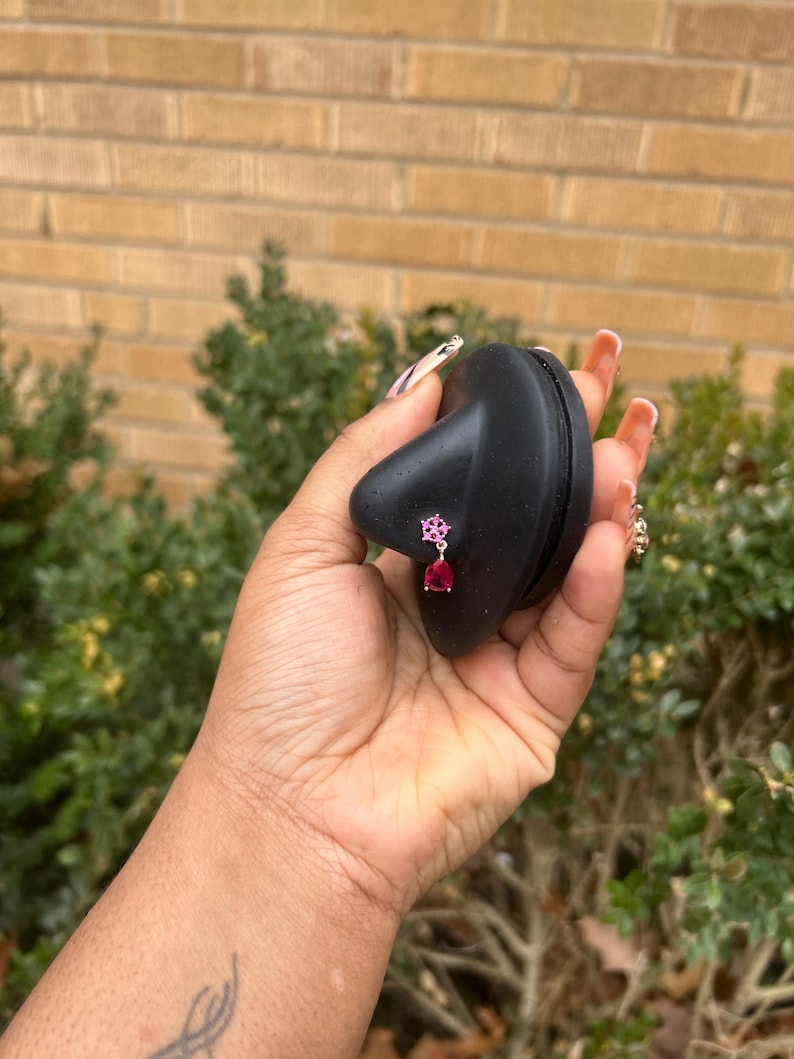 May include: A pair of pink and red gemstone earrings. The earrings are held by a black object. The earrings have a floral design with a teardrop-shaped red gemstone hanging below. The background is a brick wall and green foliage.