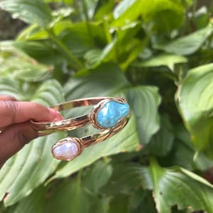 May include: A gold-toned cuff bracelet featuring two large, irregularly shaped gemstones. One gemstone is a light blue color, and the other is a creamy white. The bracelet is set against a backdrop of lush green foliage.