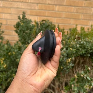 May include: A black nose-shaped display stand holding a small, dangling earring. The earring has a pink floral design and a teardrop-shaped red gem. The background features a brick wall and green foliage.